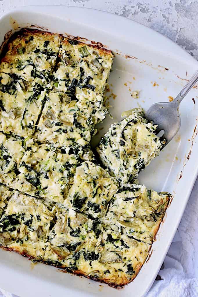 Photo of Spinach Artichoke Squares in white square baking dish cut into squares with serving fork.