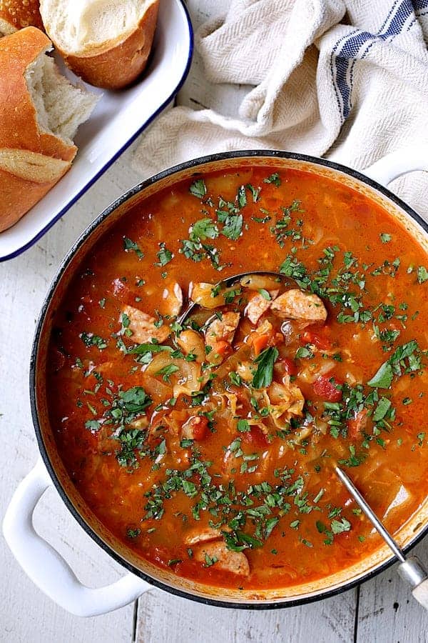 Photo of Farmhouse Cabbage Soup with Cannellini Beans and Kielbasa in white Dutch oven with ladle garnished with parsley.