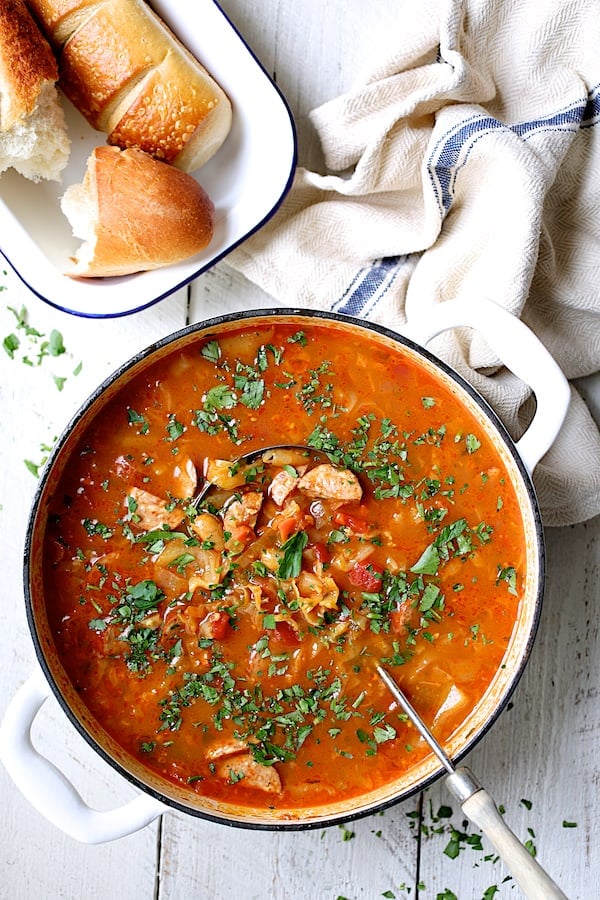 Photo of Farmhouse Cabbage Soup with Cannellini Beans and Kielbasa in white Dutch oven with bread in dish nearby.