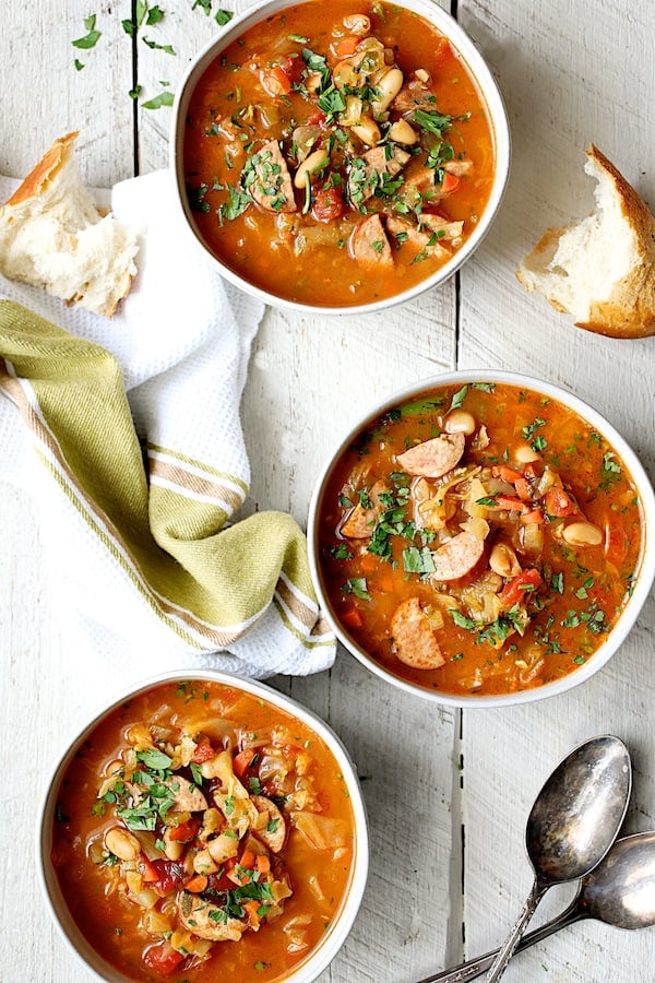 Photo of three servings of Farmhouse Cabbage Soup with Cannellini Beans and Kielbasa on white wooden background with green towel and broken bread.