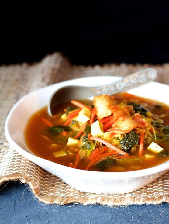 Photo of Asian Vegetable Soup with Tofu Kimchi and Rice Noodles in white bowl on brown burlap and blue background.
