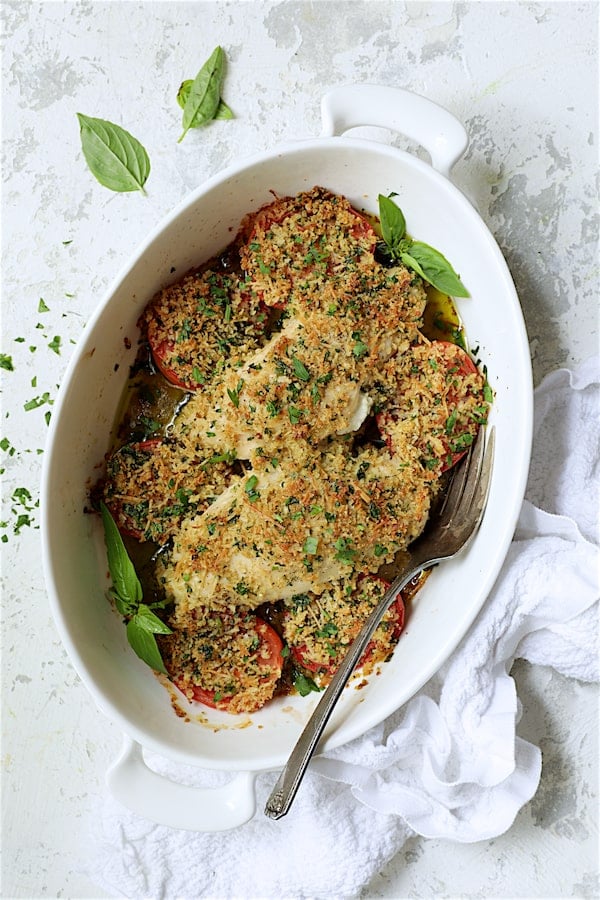 Photo of Pesto and Goat Cheese-Stuffed Chicken with Roma Tomatoes on white distressed background with white kitchen towel garnished with fresh basil.