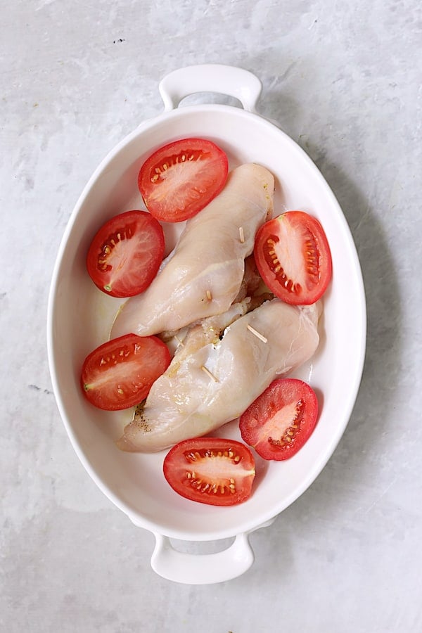 Photo of stuffed chicken breasts and plum tomatoes in white oval baking dish.