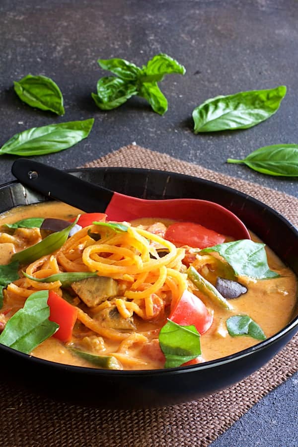 Photo of Thai Red Curry with Vegetables and Sweet Potato Noodles in black bowl with red and black porcelain spoon garnished with fresh basil.