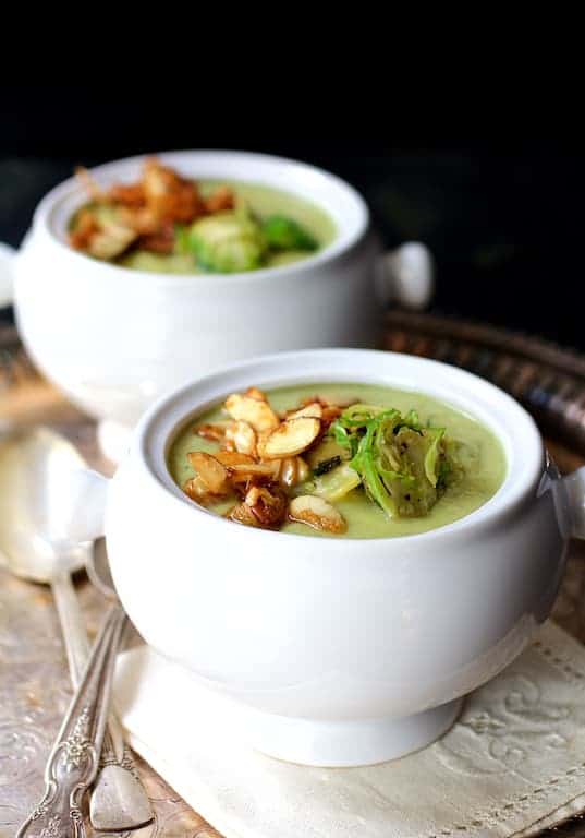 Close-up photo of a bowl of Cream of Brussels Sprouts Soup with Butter Roasted Almonds on embroidered napkin with another bowl in the background.