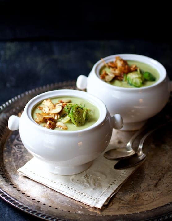 Photo of two bowls of Cream of Brussels Sprouts Soup with Butter Roasted Almonds on serving platter with one on an embroidered napkin.
