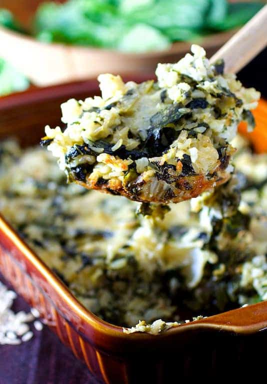 Photo of a spoonful of Cheesy Brown Rice and Greens Casserole being lifted out of brown clay baking dish.