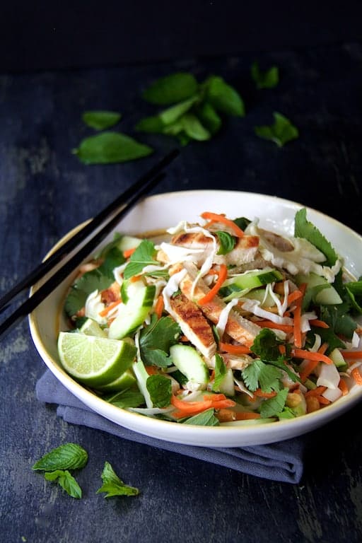 Photo of Vietnamese Chicken Salad with Spicy Lime Ginger Dressing in white bowl with black chopsticks set on blue napkin garnished with fresh mint leaves.