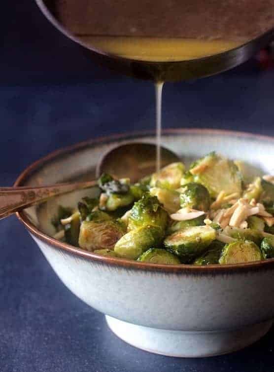 Roasted Brussels Sprouts with Browned Butter and Almonds with butter being poured over in serving bowl with serving spoon.
