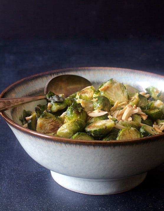 Photo of Roasted Brussels Sprouts with Browned Butter and Almonds in serving bowl with serving spoon on dark blue background.