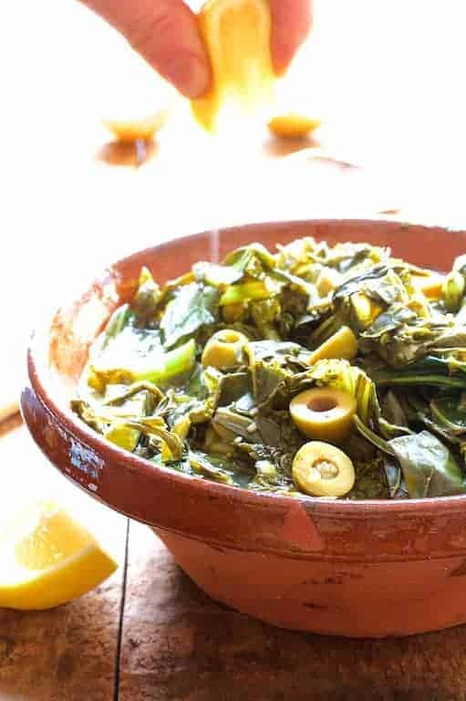 Braised Greens with Olive and Lemon in earthenware bowl with hand squeezing lemon juice on it.