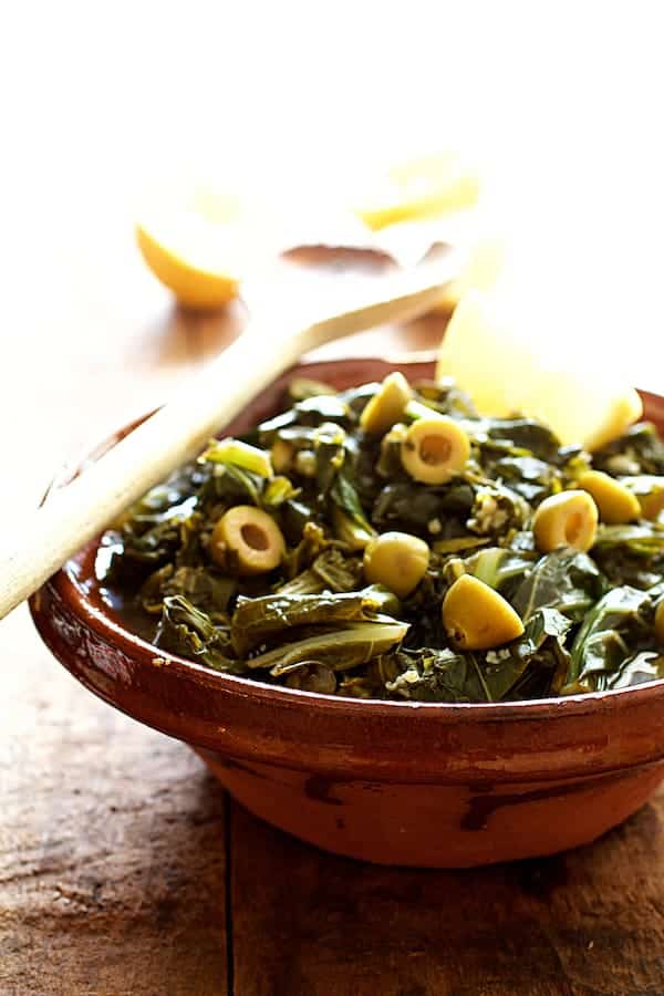 Photo of Braised Greens with Olives and Lemon in clay bowl with serving spoon garnished with lemon.
