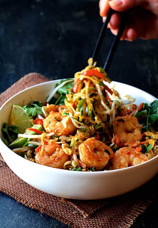 Photo of Shrimp Pad Thai with Zucchini Noodles in white bowl on brown burlap with zucchini noodles being pulled up out of the bowl with chopsticks.