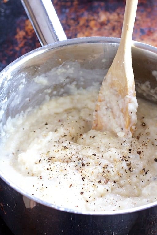 Photo of grits being cooked and stirred with wooden spoon in stainless steel saucepan.