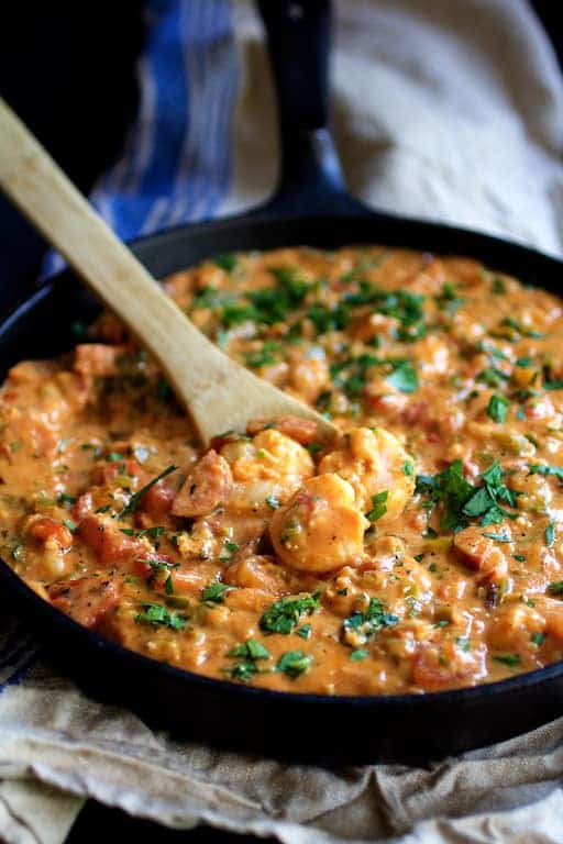 Smothered Shrimp with Andouille Sausage and Creamy Parmesan Peppercorn Grits in cast iron skillet being stirred with wooden spoon on blue striped towel.