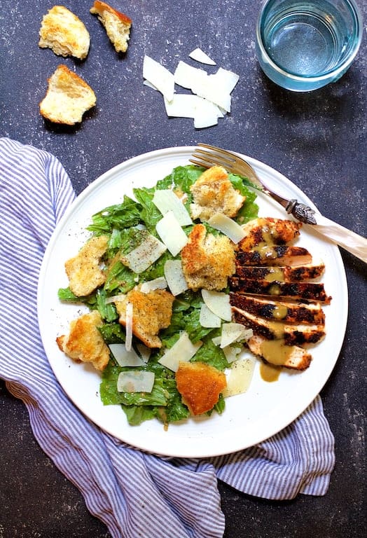 Photo of Blackened Chicken Caesar Salad with Sourdough Croutons on white plate on dark blue background with striped napkin.