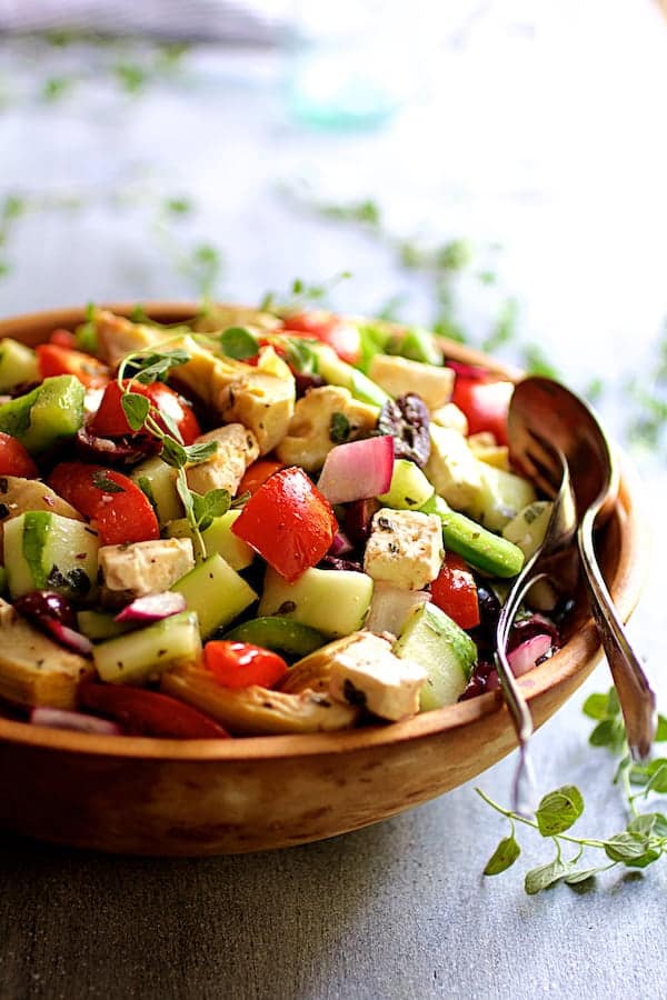 Photo of Greek Vegetable Salad with Marinated Feta Cheese in wood bowl with serving utensils garnished with fresh oregano.