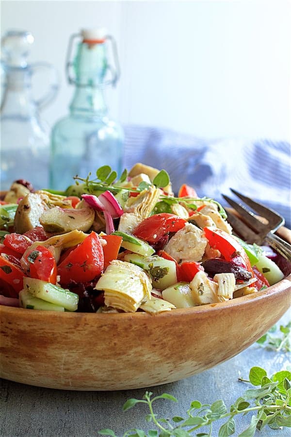 Photo of Greek Vegetable Salad with Marinated Feta Cheese in wood bowl garnished with fresh oregano with bottles in the background.