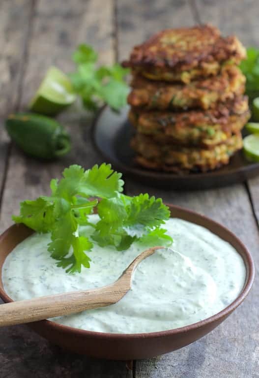 Close-up photo of sour cream in bowl with wooden spoon and fritters in the background.