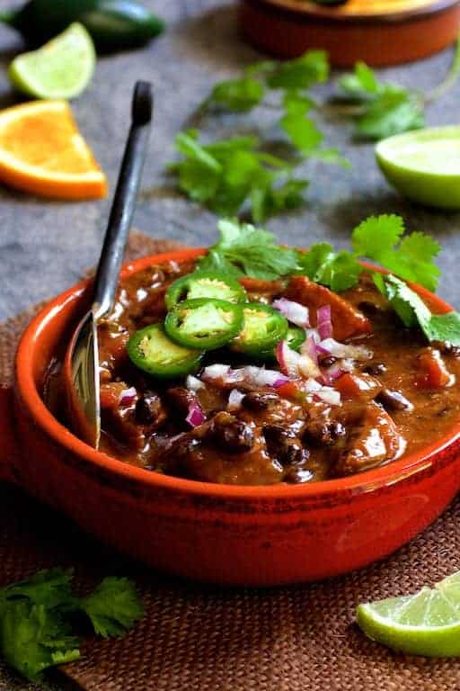 Feijoada (Brazilian Beef Stew) in red bowl garnished with jalapeno slices, cilantro and lime wedges.