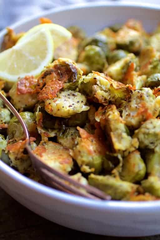 Close-up photo of Lemon Parmesan Crusted Brussels Sprouts in white bowl with serving spoon and lemon wedges.