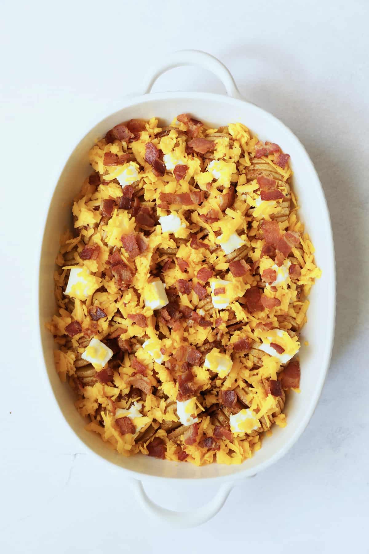 Photo of potatoes with butter, cheese and bacon in white oval baking dish ready to be baked.