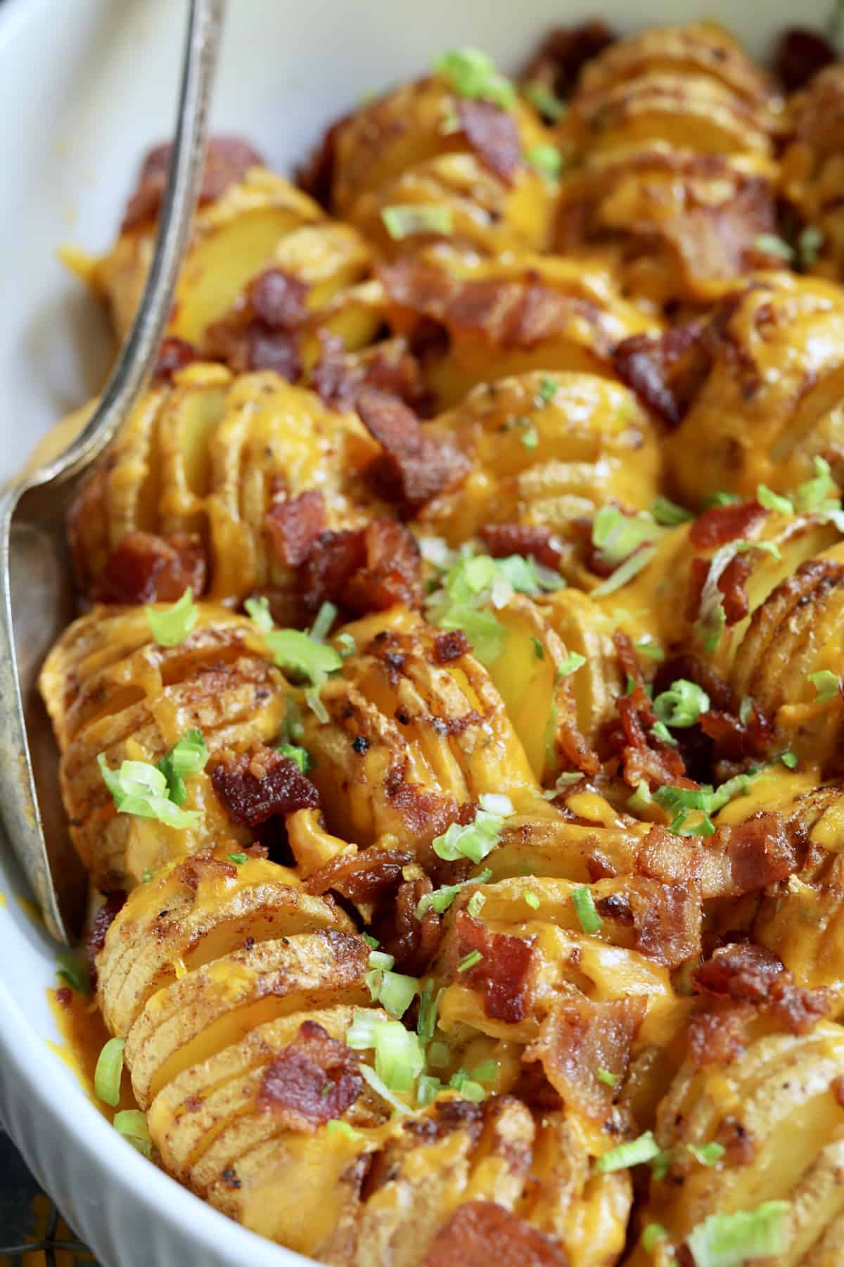 Close-up photo of Baby Hasselback Potato Bake with Cheddar and Bacon in white baking dish with serving spoon.