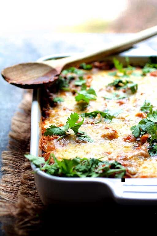 Photo of Chicken Poblano and Black Bean Tortilla Casserole in white ceramic baking dish with wooden spoon resting on the edge.