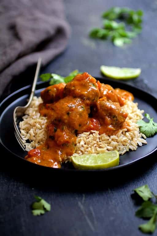 Photo of Meatballs in Spicy Curry over basmati rice on black plate garnished with lime. wedges and cilantro sprigs