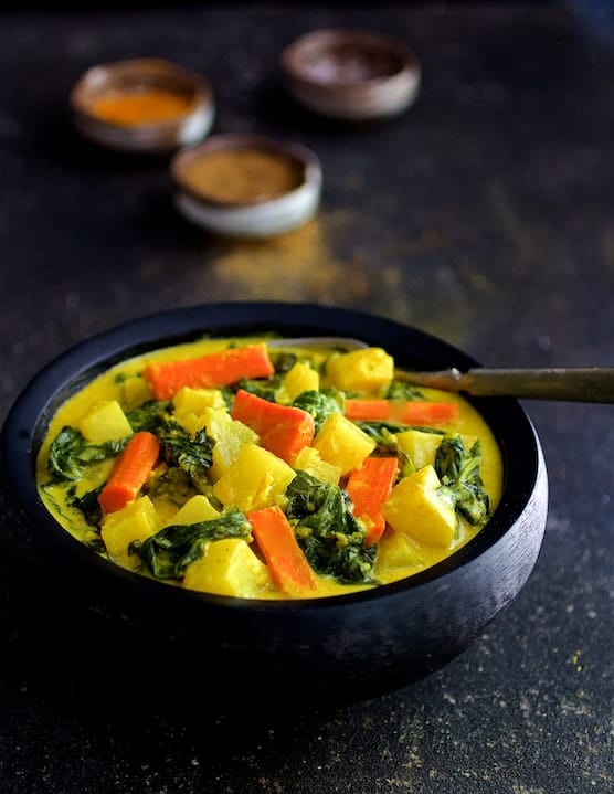 Photo of Indian Root Vegetable Curry in black bowl with small bowls of spices in the background.
