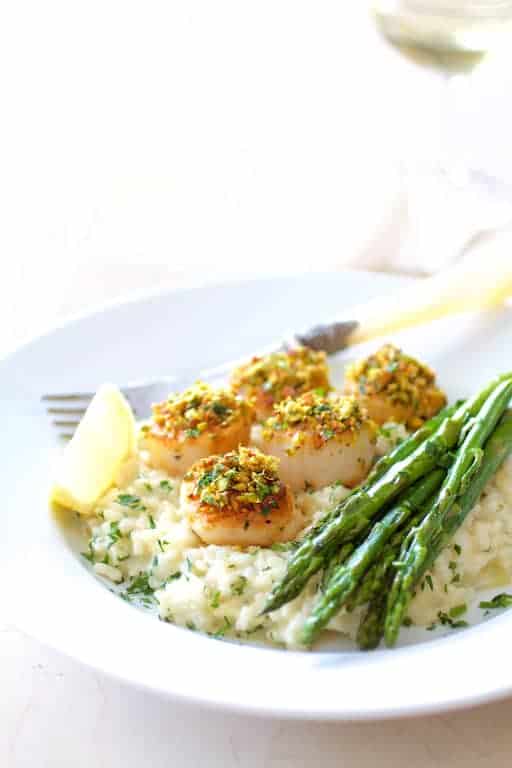 Photo of Pistachio Encrusted Sea Scallops with Champagne Risotto and Roasted Asparagus on white plate garnished with lemon wedge and wine glass in background.