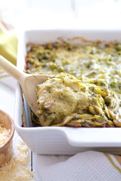 Photo of Baked Orzo with Pesto Peas Prosciutto and Mascarpone in white baking dish with some being scooped out with wooden spoon.