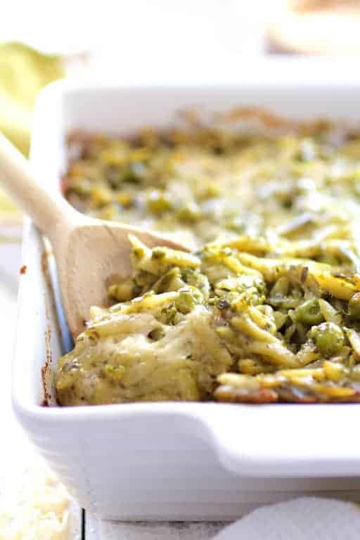 Baked Orzo with Pesto Peas Prosciutto and Mascarpone in white baking dish with wooden spoon scooping some out.