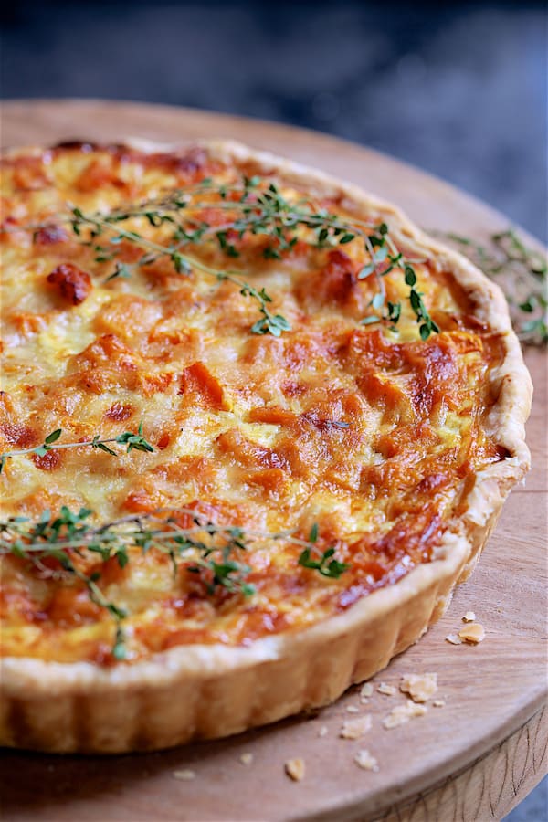 Close-up photo of whole Butternut Squash Leek and Gruyere Tart on wood stand garnished with fresh thyme.