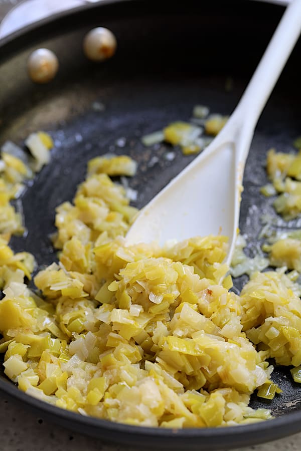 Photo of leeks being sauteed in nonstick skillet and being stirred with white spoon.