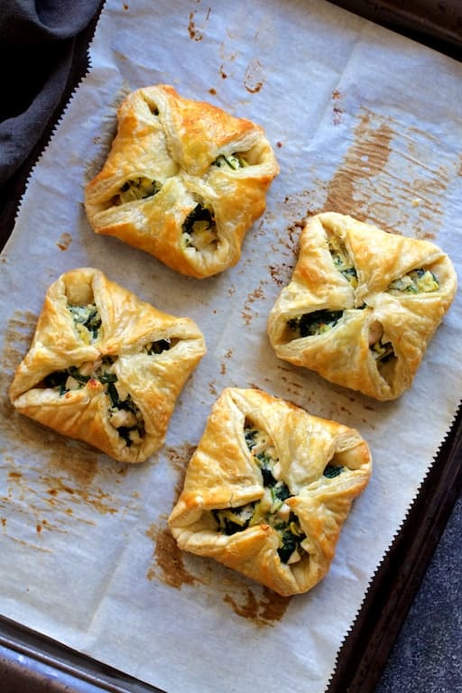 Photo of four baked Chicken, Spinach and Artichoke Puff Pastry Parcels on parchment paper-lined baking sheet.