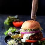Lentil Walnut Veggie Burgers with Avocado Tzatziki with red onion, tomato, alfalfa sprouts and avocado on black plate.