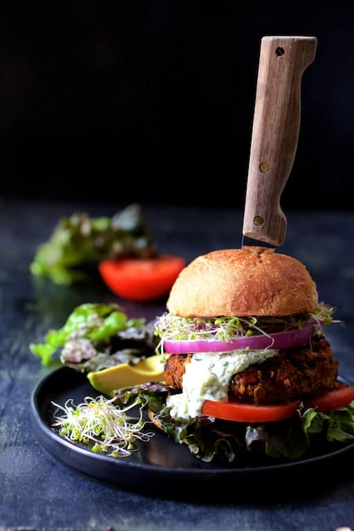 Lentil Walnut Veggie Burgers with Avocado Tzatziki with red onion, tomato, alfalfa sprouts and avocado on black plate.