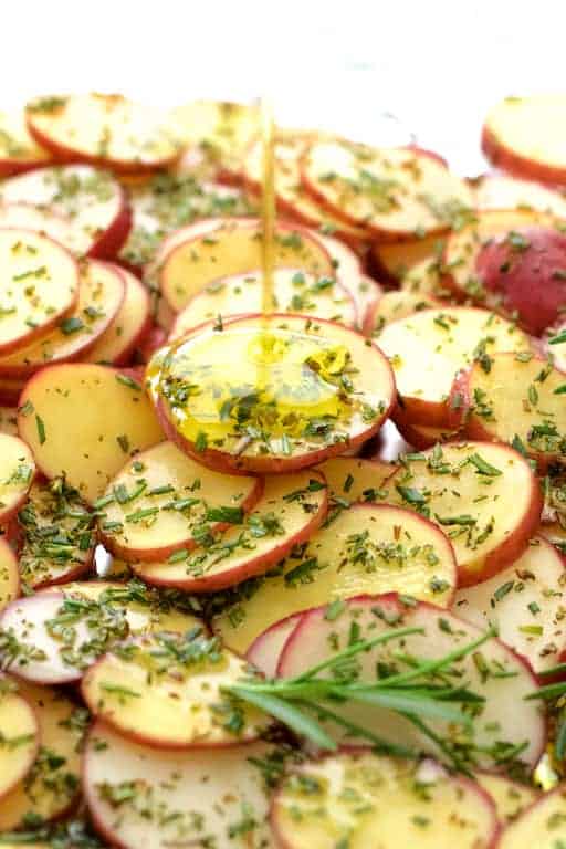 Photo of thinly sliced red potatoes sprinkled with rosemary being drizzled with olive oil.