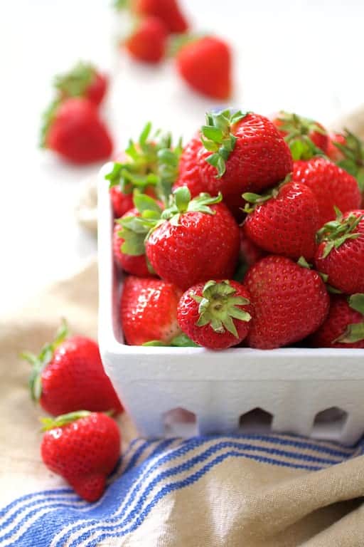 Photo of fresh strawberries in white berry basket on blue-striped towel.