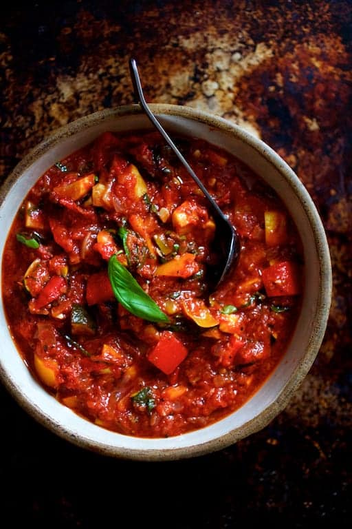 Photo of ratatouille in clay bowl with black serving spoon and garnished with fresh basil.
