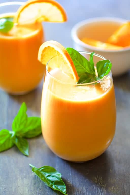 Chilled Cantaloupe Soup glasses with basil leaves on the surface and orange wedges in small white bowl in the background.