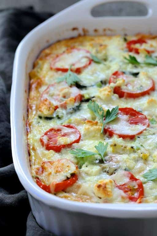 Photo of baked Four Cheese Strata with Zucchini and Tomatoes in white baking dish garnished with fresh parsley.