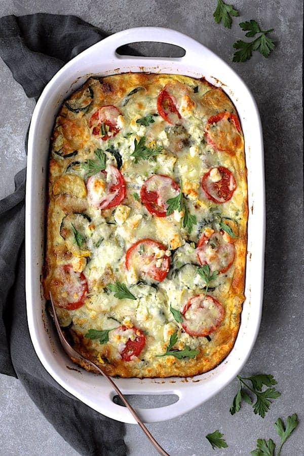 Photo of Four Cheese Strata with Zucchini and Tomatoes in white baking dish on gray background garnished with fresh parsley.