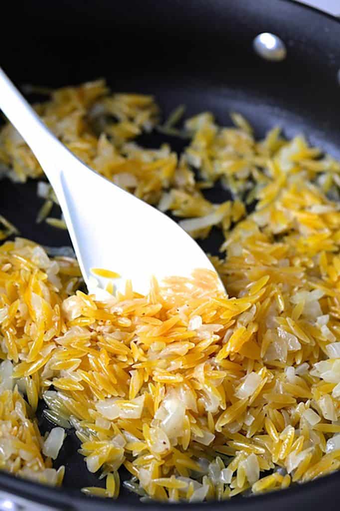 Photo of orzo being stirred in hot fat in saute pan with white spoon.