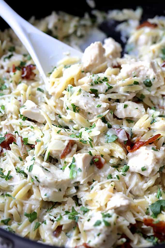 Close-up photo of Creamy One Pot Chicken and Orzo Risotto with Bacon being stirred in saute pan with white spoon.