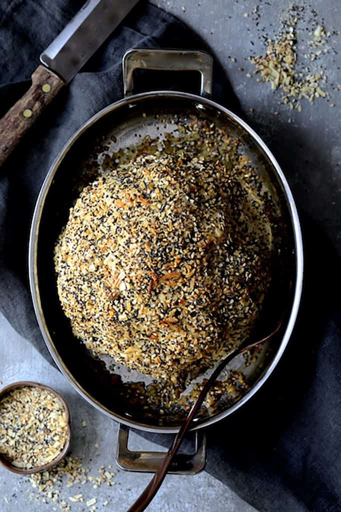 Photo of Everything Bagel Spice Whole Roasted Cauliflower in roasting pan on dark gray towel with everything bagel spice scattered on the surface.