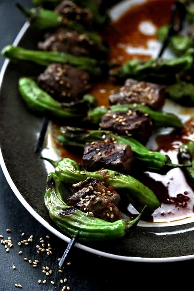 Close-up photo of Japanese Beef and Shishito Pepper Skewers on platter with sesame seeds scattered on surface.