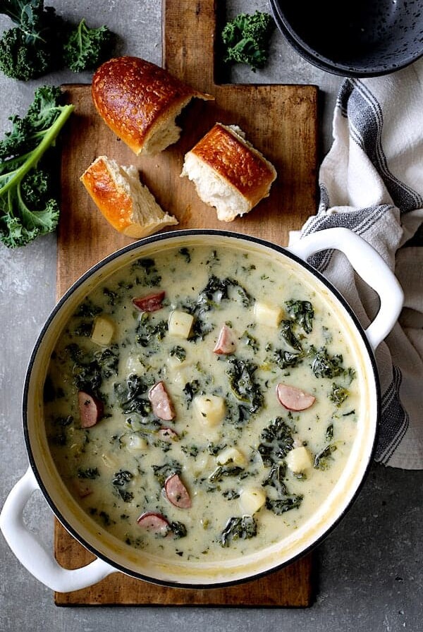 Creamy Potato Kielbasa and Kale Soup in white Dutch oven with bread and fresh kale scattered about.