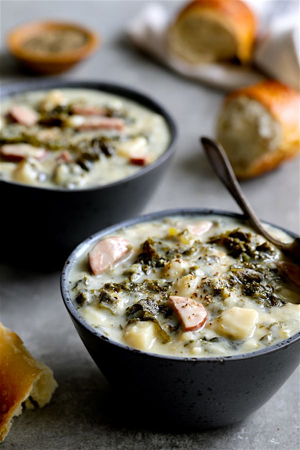 Photo of two servings of Creamy Potato Kielbasa and Kale Soup in gray bowls with bread scattered about.